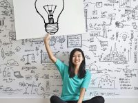 pexels-photo-3758105-3758105 Young woman holds a light bulb drawing against a creative brainstorming wall.